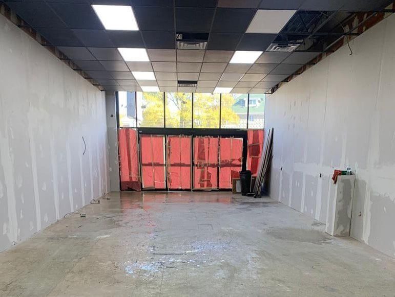 A wide-angle view of a commercial space under renovation, with exposed concrete floors, new drywall on the walls, and a large front window partially covered by red construction paper.